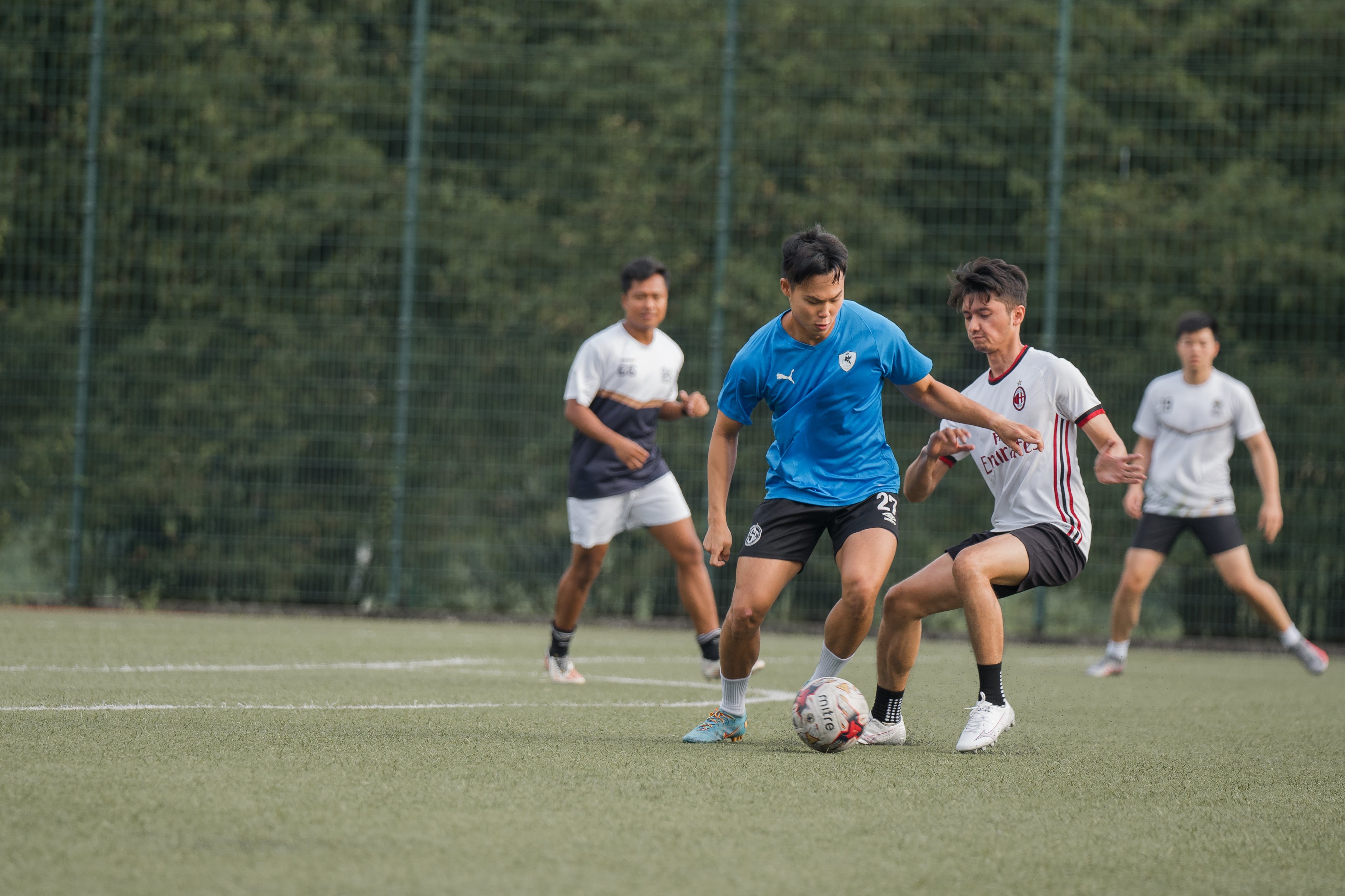 Two soccer players contest the ball on a green turf field, with two teammates visible in the background.
